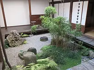 A garden courtyard with an en about 30 centimetres (12&nbsp;in) above the pale gravel, feature stones, a rain chain, and a planting of bamboo, ferns, grasses, and a creeping plant with small round leaves.