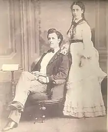Black and white of a middle-aged man and woman. The man has dark hair, is seated in an arm chair, wearing a dark-colored suit jacket, light-colored pants, white dress shirt and black bowtie. The woman is standing behind and to the side of the chair, wearing a full-length white dress with a dark-colored belt, and dark hair braided into two ponytails.