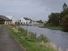 A view along a canal with a lock and buildings in the background. Reeds and other water plants line the canal.
