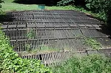 A horizontal wooden rack roughly 30 feet (9.1&nbsp;m) wide and 30 feet (9.1&nbsp;m) long lies in flat, grassy area.