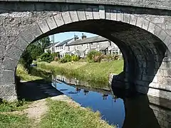 Close up of grey stone arched bridge over narrow canal with single sided towpath on a sunny day reflecing as blue on the still water