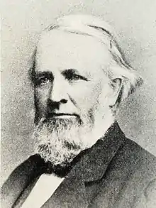 Head and shoulders photograph of a man in an overcoat. He has a full beard with sideburns and has his white hair parted at the his right with it being of enough length to be tucked a few inches behind his left ear. He wears a serious expression and is looking to the left.