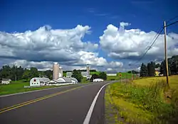 Country road with blue sky and clouds rolling overhead. There is a farm on the left and a sign indicating an intersection on the right.