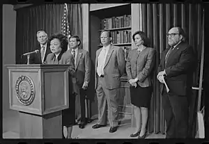 A photograph of a woman standing at a podium, behind whom are a group of four men and one woman standing against a draped wall with a bookcase