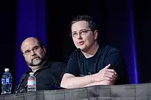 An American man with graying black hair sits behind a stand speaking to a crowd through a microphone.