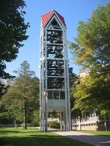 A three-dimensional vertical structure perhaps 70 feet (21&nbsp;m) tall and 10 feet (3.0&nbsp;m) wide by 10 feet (3.0&nbsp;m) long is filled with tiers of large metal bells. The structure, standing in front of a large institutional building in a park-like setting, is supported by several metal legs. A sidewalk passes through the structure, which is open at the bottom to a height of perhaps 10 feet (3.0&nbsp;m).