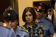 A serious-looking woman with a brown bob haircut and plaid shirt, is led into a courtroom between two uniformed officers.