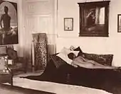 Naked youth poses prone in the Villa Lysis of Capri, where Nino Cesarini lived with his protector and lover, the Baron Jacques d'Adelswärd-Fersen. The photograph was taken by Wilhelm von Plüschow and the portrait of Cesarini is visible on the wall.