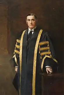 Three quarter length standing portrait, the subject looking directly at the viewer, while turned slightly to left. One hand rests on a low wooden table and he wears gowns which are black with a gold trim and details. Standing against a brown background.