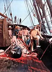 A group of men on the deck of a ship surround the remains of a whale, with much blood and blubber in evidence. The figure on the right, the prince, is wearing a tailored jacket and smart hat. The others are in seamen's jerseys.
