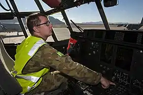 Bespectacled man in camouflage uniform with fluorescent jacket in cockpit of military aircraft