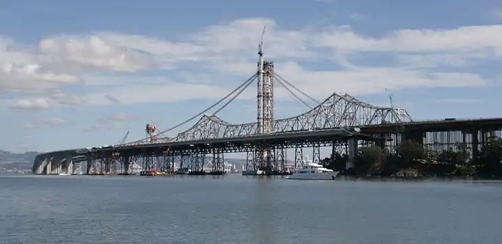 October 1, 2011: In the distance, the Left Coast Lifter is placing the last of the main span deck segments. Two additional short segments will join the main span to the curved skyway extension. The main suspension cables will follow the curves outlined by the recently installed catwalks. Ten holdback cables to the right of the tower (below the catwalks) preload the tower, bending it 17 inches (430&nbsp;mm) west against the forces to be imposed by the main cable when the bridge is complete, allowing the tower to be vertical when the holdbacks are removed. Subsequent to this image the traveler support cabling and cabling supports were installed, and all of the main cable strands have been placed and compacted, the suspender cables hung, attached, and tensioned.