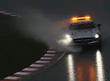 A large amount of water is sprayed from the tyres of a silver sports car with orange hazard lights on the roof.