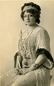 A white woman with curly dark hair piled up loosely and wrapped with a scarf; she is seated and wearing a light-colored gown with extensive beading